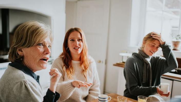 Three females are sitting around a wooden table within a kitchen. They are drinking tea there is two tea mugs and two juice glasses on the table. The kitchen is white with wooden counter top. There is a white cooker, white door and white window in the kitchen. 