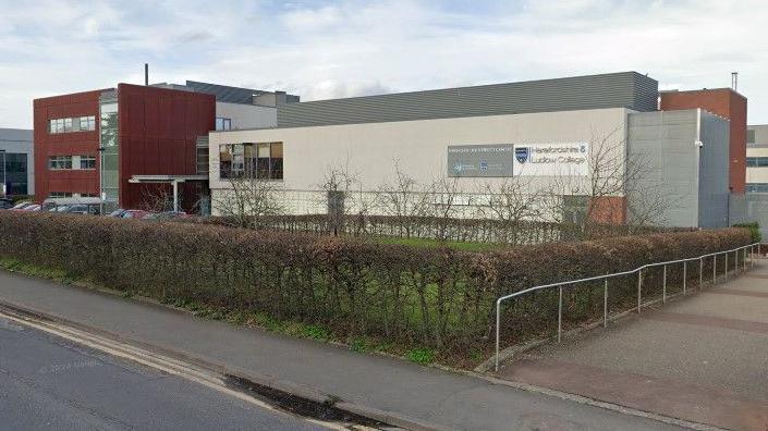 The front of the college buildings seen from the street. A long white building forms the majority of the frontage, with a shorter, taller dark red block at the end. A neat hedge surrounds the front of the grounds.