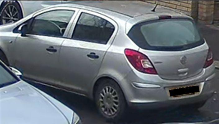 A CCTV photo of a silver Vauxhall Corsa driving down a road past two other parked cars.
