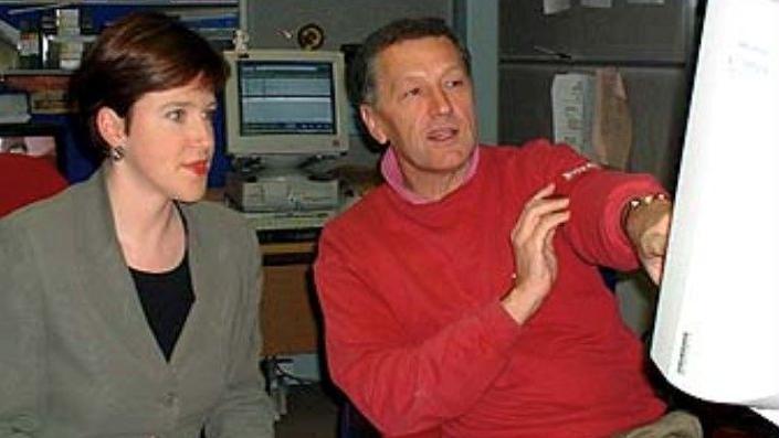 Susie Fowler-Watt, wearing a green-grey jacket and black round-neck T-shirt looks at a computer screen with Stewart White, who is wearing a red jumper and khaki trousers