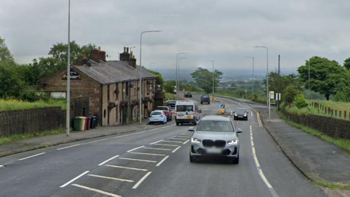 Car driving along Chorley Old Road, which is lined by trees, green fields and an old terrace of buildings