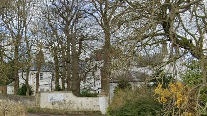 Howlish Hall care home is a white building tucked behind a row of trees and a white wall.
