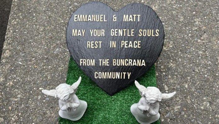 A heart shaped sign saying: 'Emmanuel & Matt May Your Gentle Souls Rest In Peace From The Buncrana Community' with two angel statues next to it.