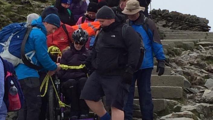 Woman in wheelchair climbing a mountain with help of others