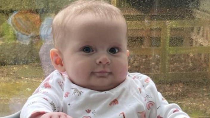 Penelope Greathead, a baby girl wearing white patterned baby clothes. She is sitting against the backdrop of a rainy window.