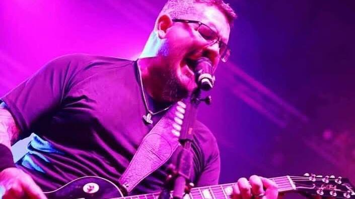 A man with glasses on and a black T-shirt. He has a guitar in his hands and is singing.