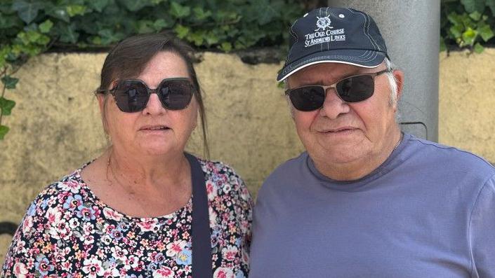 Maggie Wright, who has tied back brown hair and her husband Colin, who wears a black and white baseball cap, stand side-by-side in front of a wall in Woking town centre, wearing sunglasses.