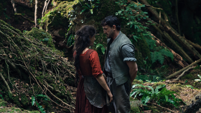 A still of the film Hamnet, showing Jessie Buckley as Agnes and Paul Mescal as William Shakespeare. The pair stand in period dress very close together in the middle of a forrest.