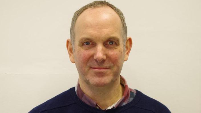 Architect Ian Thorp wearing a blue sweater and red shirt smiles for the camera. 