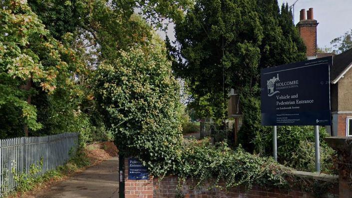 A blue school sign is on the right, adjacent to a path leading to a school. There is a small brick pillar next to the path which is covered in leaves. There are many trees in the background leaning over the path