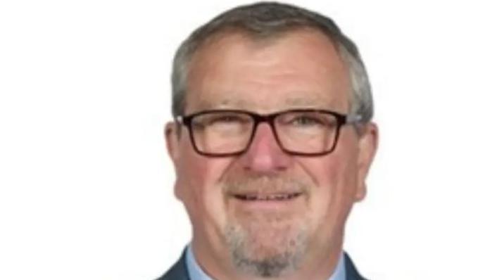 A headshot of a man with grey hair and grey beard. He has glasses and a shirt collar and jacket can just about be seen