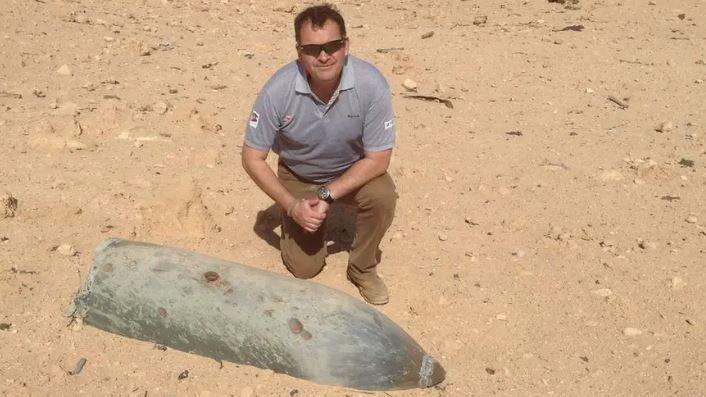 Ben Remfrey wearing a grey polo shirt and brown trousers kneeling down on sandy ground next to the shell of an explosive.