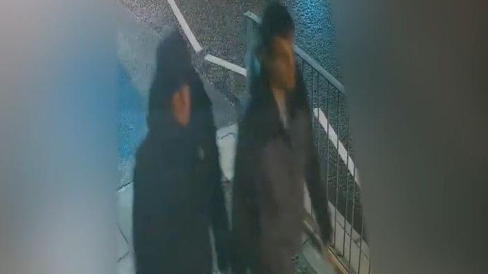 A still from CCTV footage of two white men, one wearing a black coat with the hood pulled up, and the other who has short, dark hair and wears a two-tone grey and black zip-up jacket, walking on a pavement