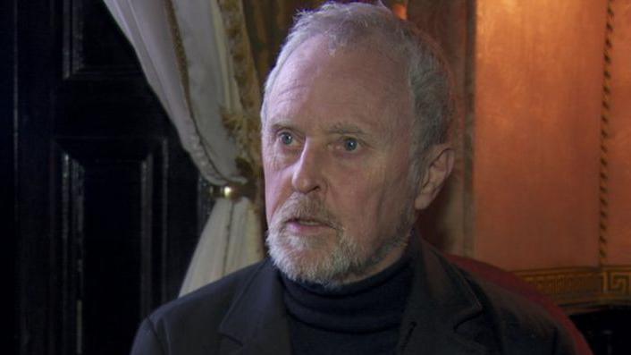 Bill Wolsey - A man with grey hair and beard sits in front of a wooden window cover, curtains and orange wall. He is wearing a dark blazer and a high neck jumper.