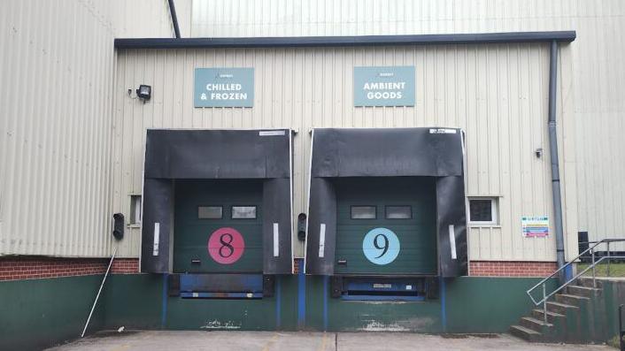 The side of a warehouse with two loading bay gates. The two doors have eight and nine written on them with signs above saying, "Chilled and Frozen" and "Ambient Goods". The doors are green.