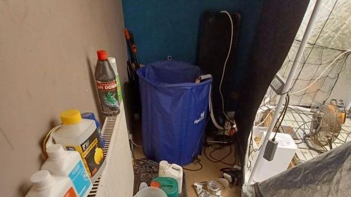 Bottles of chemicals are standing on a radiator and there is cannabis farm equipment everywhere including a blue bucket and lots of wires.