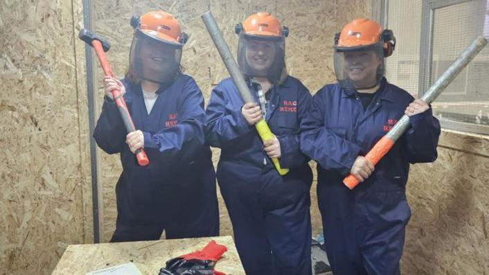 Three women holding axes and metal pipes smiling to camera wearing overalls and orange helmets to camera in a small room surrounded by chipboard