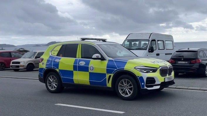 A side view of a yellow and blue large Guernsey police BMW vehicle. It is driving along a Guernsey coast road. Other cars are visible behind it, parked along a sea wall. The sea is in the background and other islands are visible in the distance. 