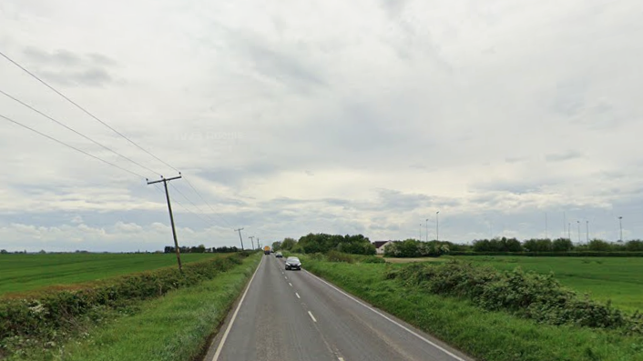 A quiet country going through open green fields, bordered by hedgerows and distant trees. Power lines trace the roadside, and a few cars travel along the tarmac under a cloudy, grey sky.