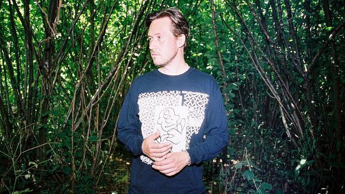 DJ Joy Orbison standing against a woodland backdrop. He is wearing a blue and white sweatshirt