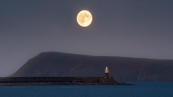 A lighthouse next to the water with a mountain in the background and the supermoon above it 