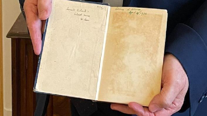 A man's hands hold the book open on the first page where there is a hand-written inscription. The book cover is blue and the pages have yellowed due to the book's age