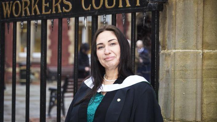 A woman wearing a navy blue and white gown wearing a teal lace dress. She has long black hair and is smiling. She stands in front of a large gate.