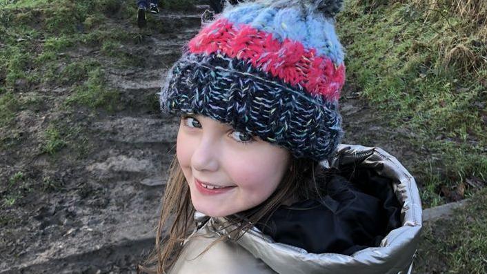 A little girl smiling wearing a black pink and white beanie hat in a silver puffer jacket