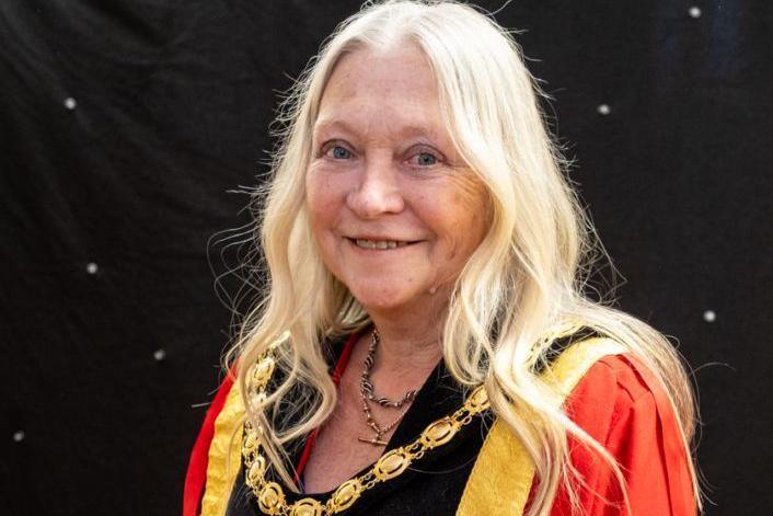 A woman with long light blonde hair, wearing a read mayoral robe and gold chain around her neck, stands and smiles in front of a black sheet with white spots.