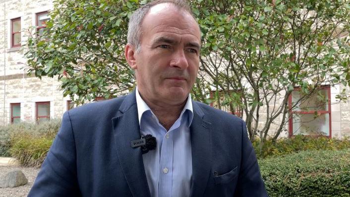 Alfred Cannan, who is wearing a dark blue suit with a white shirt.
He is standing outside the Noble's Hospital building and a tree and green bushes can be seen in the background.