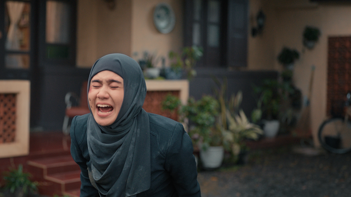A still from the movie, which shows Norma in dark blue tudong screaming outside a house.