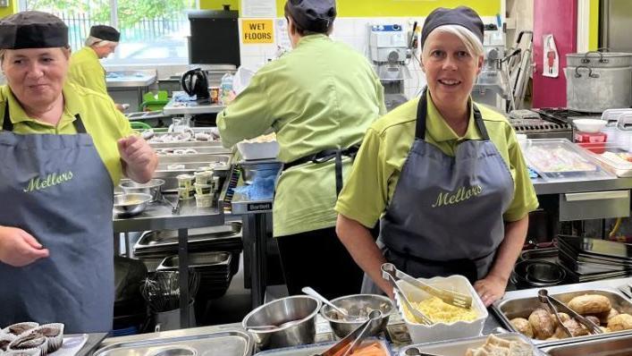 Dinnertime at Whale Hill Primary School, Eston. Lunch staff in hats and grey aprons standing in from of silver hot plates of food including baked potatoes, baked beans, crusty bread, carrots and cucumber.