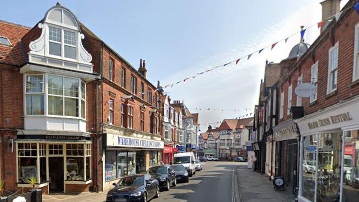 Church Street in Sheringham. It is a narrow road with shops on either side. Cars are parked on the left. Bunting is extended between the buildings below a blue sky.