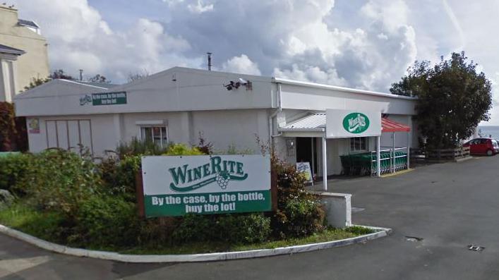 The exterior of Winerite, which is a large white building with a car park in front and a white and green sign that reads WineRite - By the case, by the bottle, buy the lot.