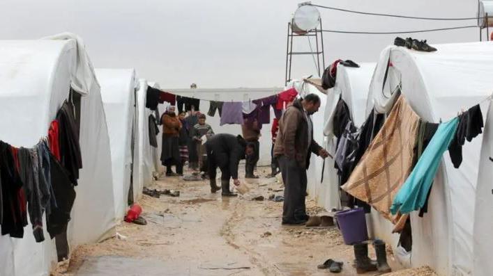 People stand near a row of white tents . They have lines with multicoloured clothing hanging from them. a child can be seen in the background.