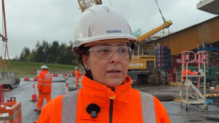 Rosario Barcena at the site where the Clifton Bridge is being built. She is wearing a white hard hat, safety goggles and orange high-viz jacket. She is looking in the distance while being interviewed by someone out of shot. She has a microphone pinned to the side of her jacket. Behind her are heavy plant machinery and workmen in high-viz clothing working around the steel structure of the new bridge.