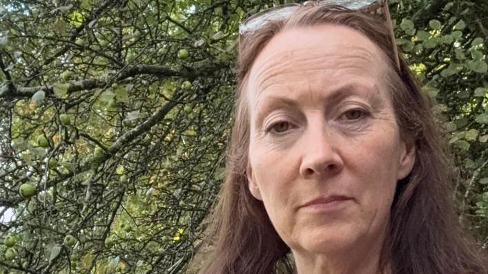 Helen, a small business owner, stands by a tree with her long brown hair blowing in the wind.
