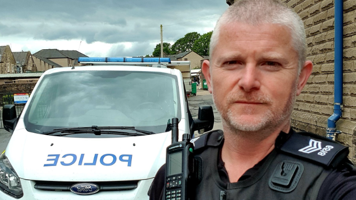 Sgt Kev Day, in his black police uniform and radio, poses in front of his patrol car