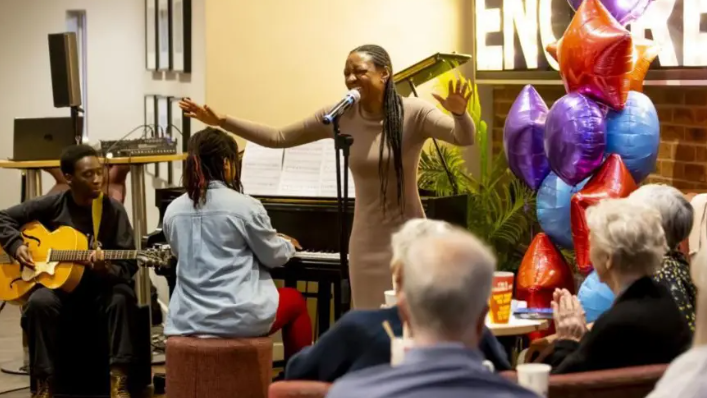 A woman in a brown dress performing into a microphone to a crowd. There are colourful balloons behind her which are purple, blue and red. There is a pianist who is wearing red leggings and a blue top, and a guitarist behind her.