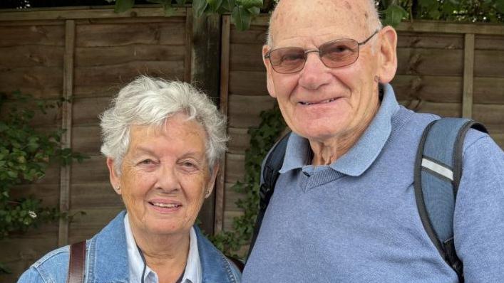 The image shows a couple.
The lady on the left is wearing a denim jacket and has short grey hair. 
The man on the right is wearing a blue jumper with a blue rucksack on his shoulders. he is wearing tinted glasses. 
Both are stood infront of a wooden fence