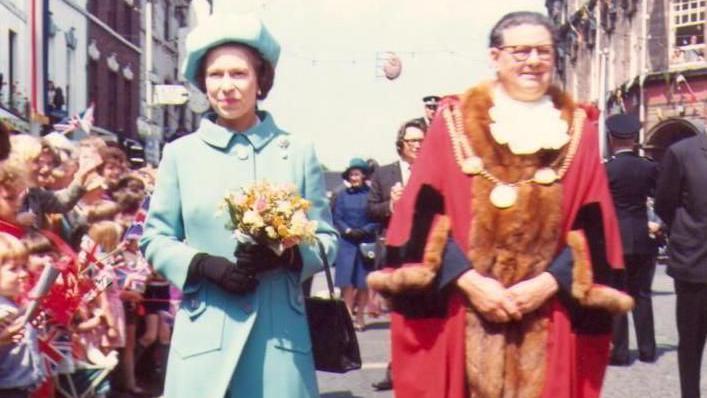 A faded colour picture of Queen Elizabeth in a turquoise dress and hat, carrying a bouquet of flowers, walking with dignitaries as she is greeted by crowds.