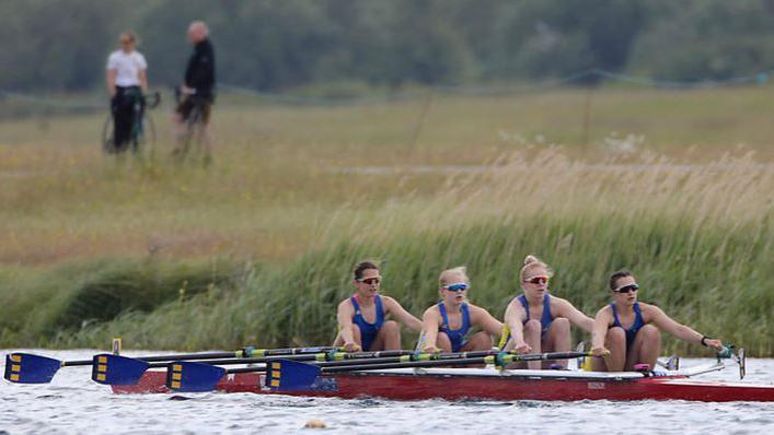 Four women rowing in a boat, wearing blue rowing kit and sunglasses. They are rowing in a river with a grassy bank in the background. 