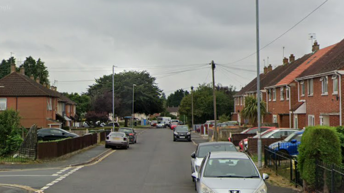 Red brick houses either side of a suburban road, with a number of cars parked on either side of the carriageway, and a cars also parked on driveways