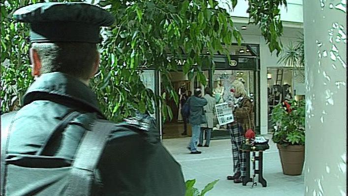 A polie officer in dark green uniform, watches on with his back to the camera, as a clown in a tartan blazer and jacket holds a newspaper in the entrance of a shopping centre.