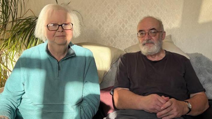 A woman in a light blue jumper sits next to her husband who is wearing a grey shirt, on a sofa. Behind them on one side are green fronds of a plant and behind them is beige patterned wallpaper