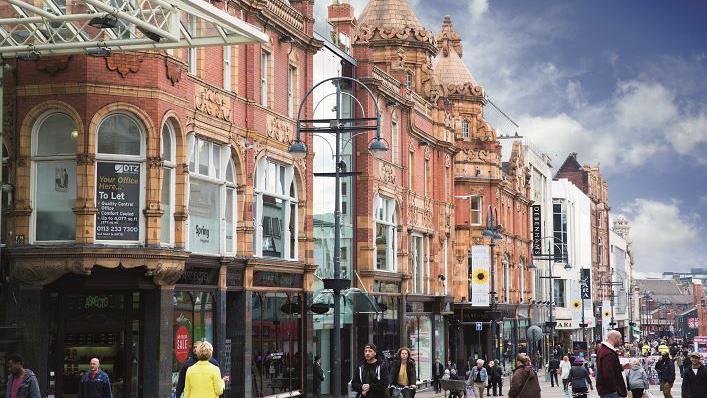 A busy shopping street in Leeds which has a row of Edwardian buildings in terracotta brick with ornate decorations and turretted roofs. 