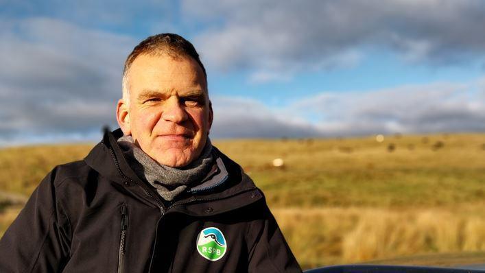 Ian Ryding is standing in the countryside, looking into the camera. He is wearing a black jacket with the "RSPB" logo on it. He has short brown hair.