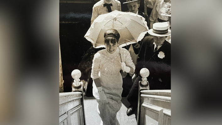An archive sepia image of Sarah Hosking as a young woman stepping onto a college barge. She is in Edwardian dress for a barge fundraiser at summer Eights week 1966. She is smiling while looking up at the camera. She is holding a parasol. Other people in period attire could be seen behind her.