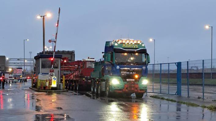 A blue and green lorry, with headlights on, being driven past a raised barrier. The lorry is carrying red machinery and driving next to a blue metal fence.  Puddles have gathered on the road. A person wearing an orange coat is standing nearby watching the lorry.