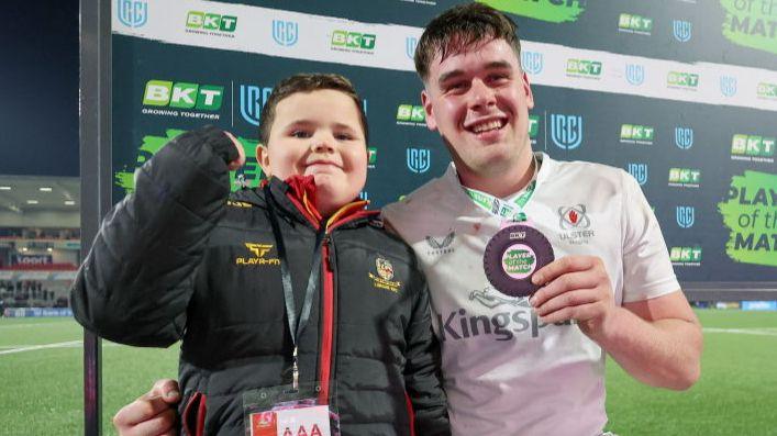 Ollie Smyth smiles with his fist in the air beside Ulster player James McNabney on the pitch at the Kingspan Stadium. Ollie has dark hair and is wearing a black and red Lurgan Rugby Club coat. James smiles holding his man of the match medal and is wearing a white Ulster Rugby kit.